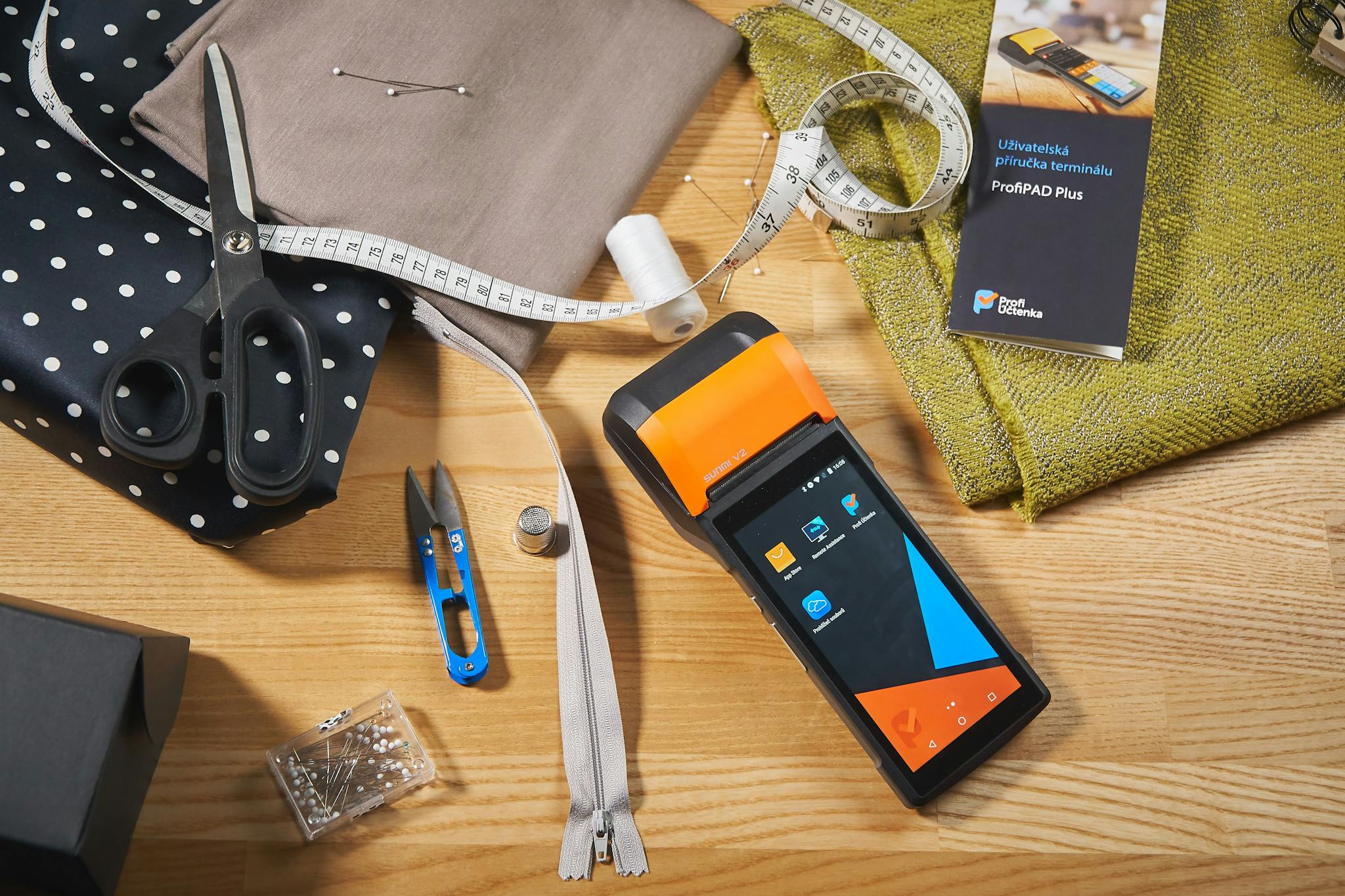 A collection of sewing tools and fabrics with a payment terminal on a wooden desk.