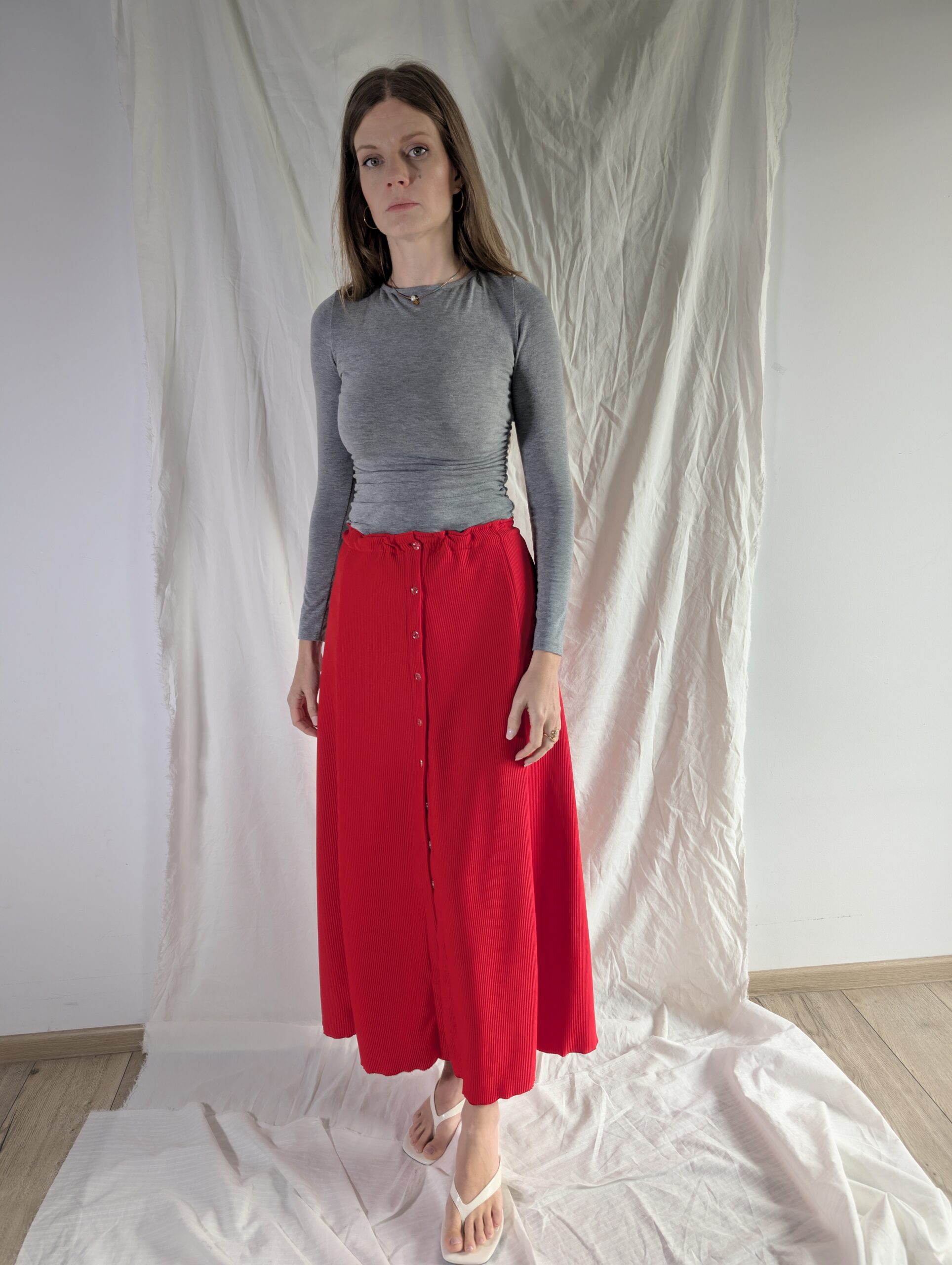 90s A-Line Skirt sewn in red ribbed knit with a button front, pleated midi length, worn with a grey long-sleeve top against a white draped backdrop