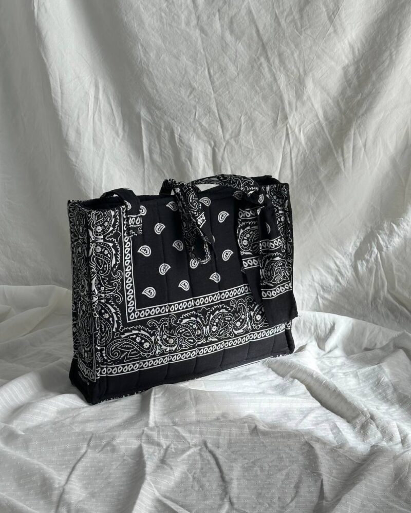 Black quilted bandana boxy bag with a white paisley print, knotted handles and vertical quilting, styled on white draped fabric